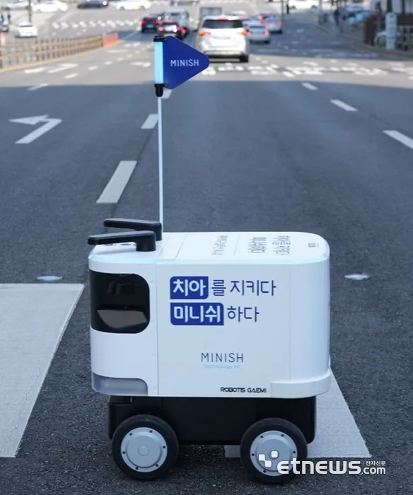 도로 위를 이동하는 MINISH 로봇 배달기기로, 흰색 몸체에 파란색 깃발이 달려 있으며 '치아를 지키다 미니쉬 하다' 문구가 적혀있다.