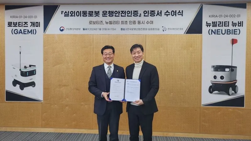두 남성이 실외이동로봇 운행안전인증서 수여식을 배경으로 인증서를 함께 들고 서 있음.