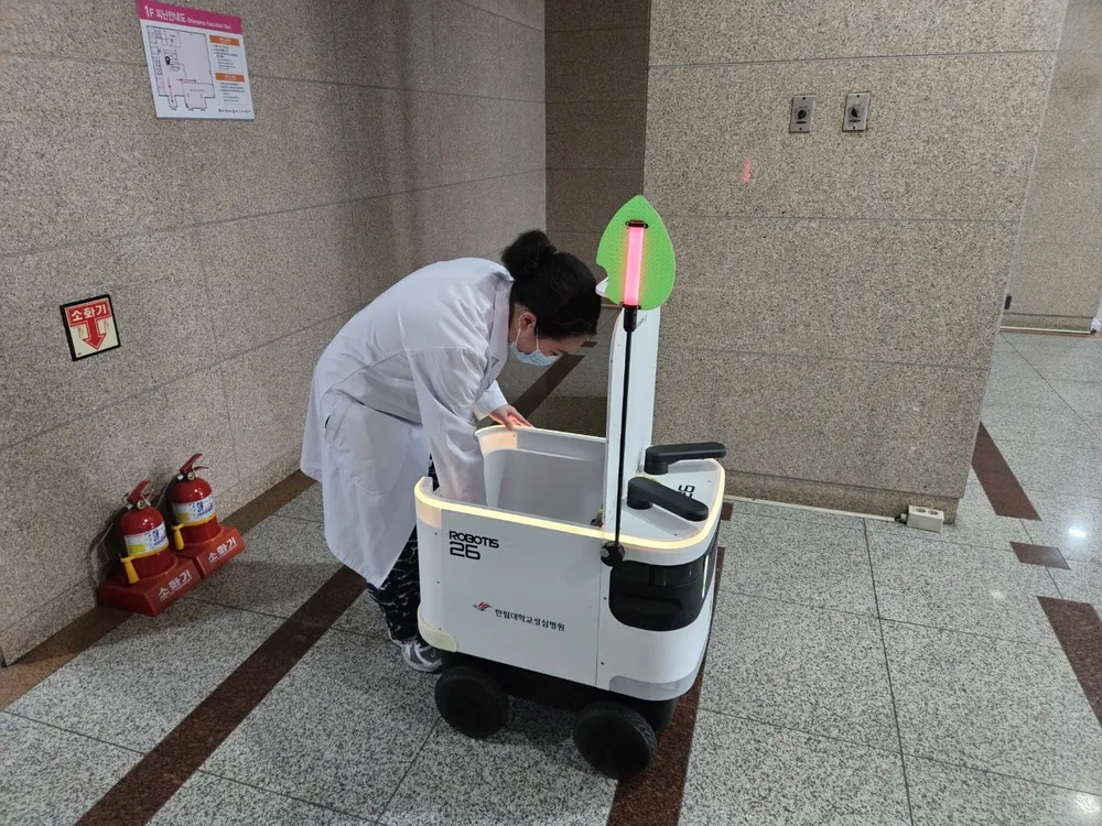 병원 복도에서 흰 가운을 입은 사람이 물품 운반 로봇에서 물품을 꺼내고 있음