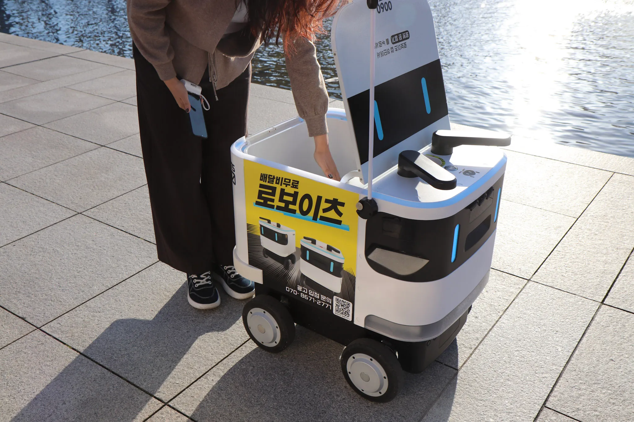 사람이 스마트폰을 들고 자동배달 로봇의 열린 칸에 손을 넣고 있는 장면.