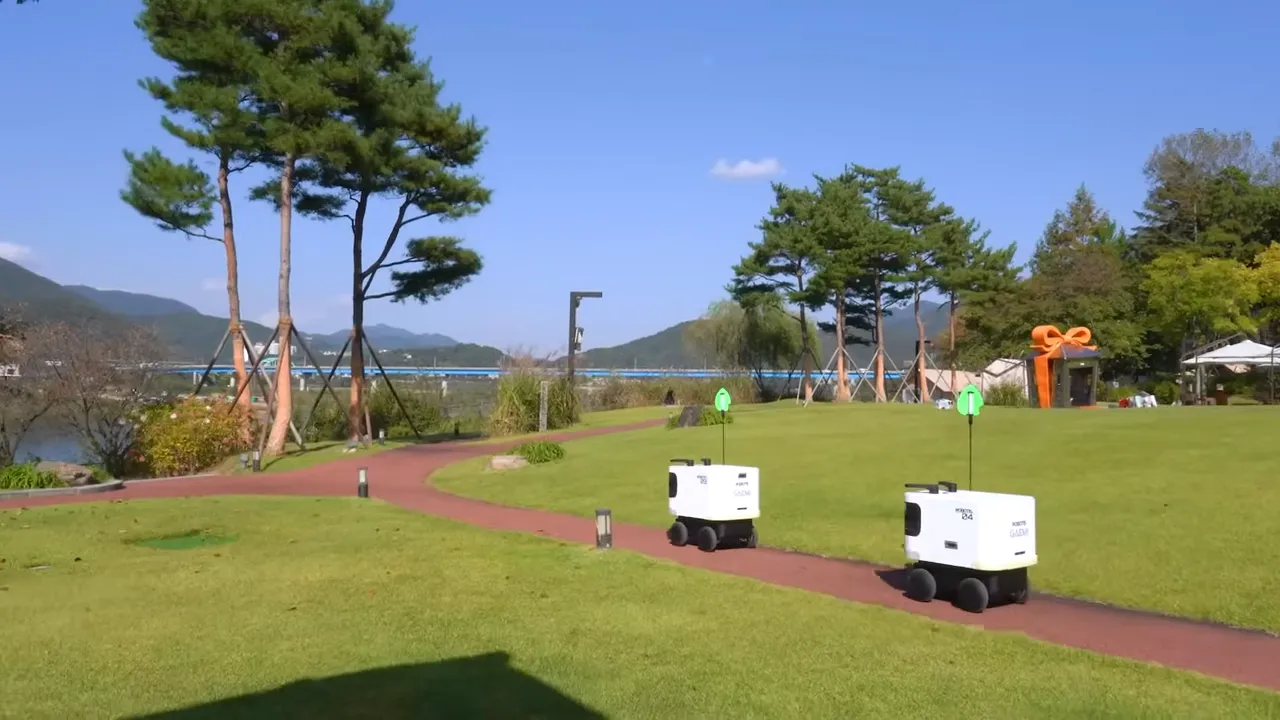푸른 하늘 아래 잔디밭 길 위를 두 대의 흰색 야외 서비스 로봇이 이동하고 있음.