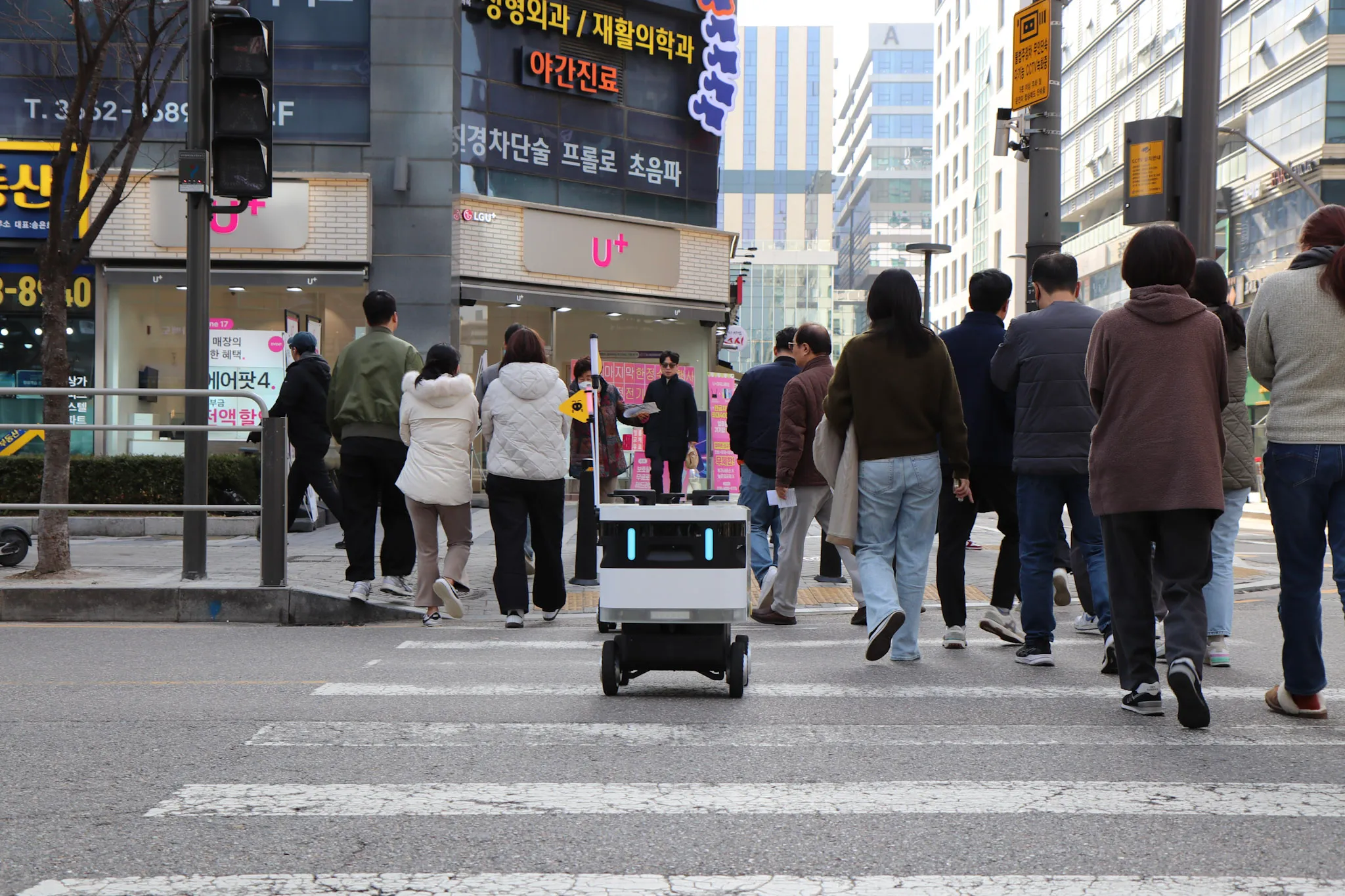 사람들과 함께 횡단보도를 건너는 배달 로봇이 도시 거리의 상점 앞을 지나가고 있다.