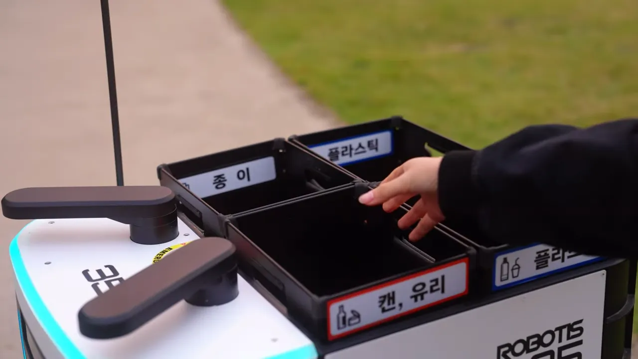 로봇 앞에 캔, 유리, 종이, 플라스틱 분리수거함과 손이 가까이 있는 모습.