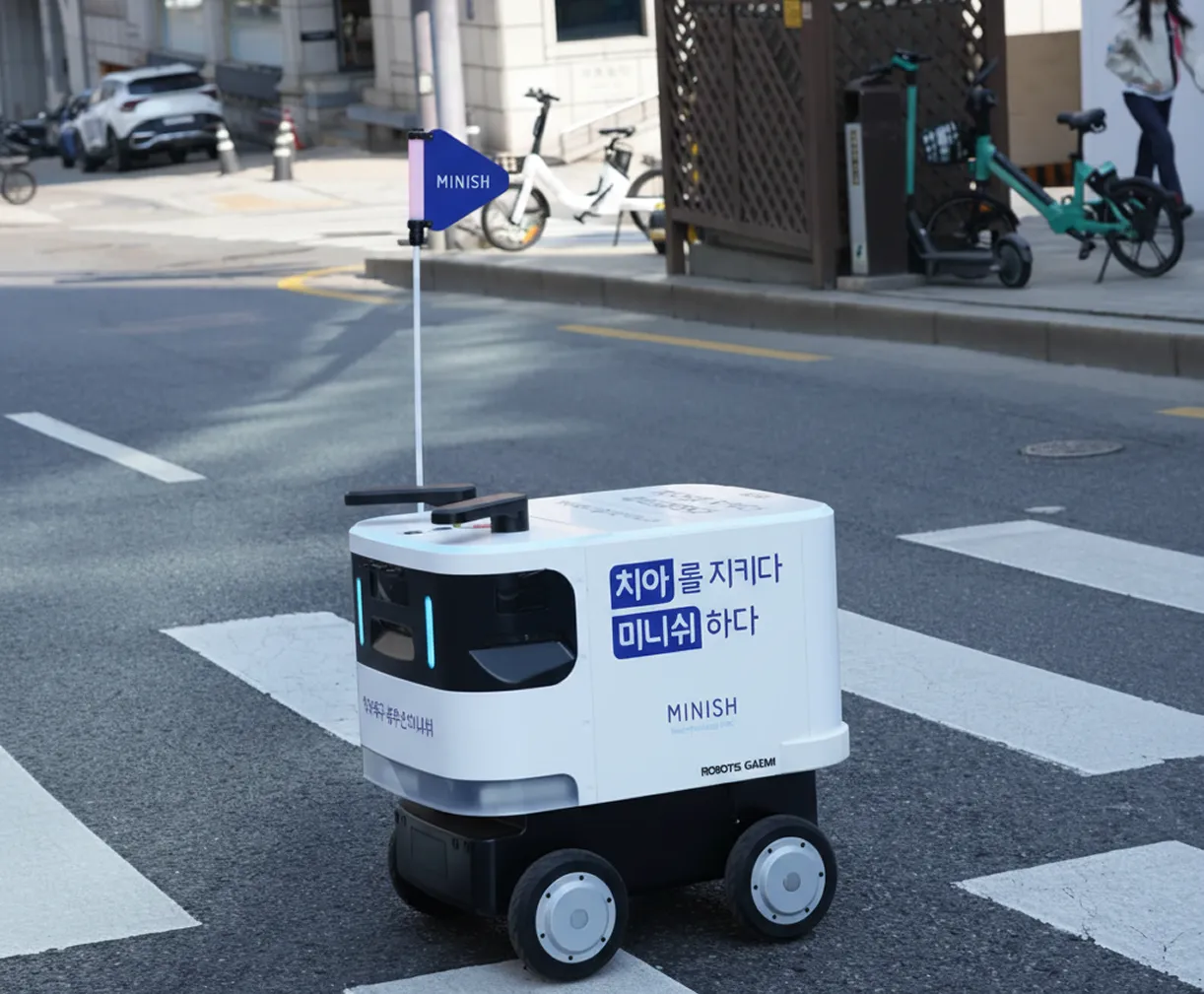 도로 위 횡단보도에 서 있는 배달 로봇 미니쉬, 흰색 본체에 파란 깃발이 달려 있음.